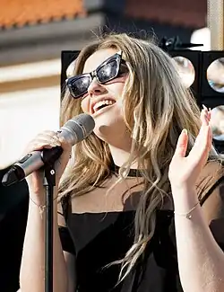 Young woman with long wavy hair wearing black dress and sunglasses singing at a microphone