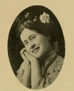 A smiling white woman with her cheek resting on clasped hands, and a rose in her dark hair
