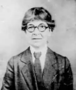 A middle-aged white woman with short greying hair, wearing round eyeglasses and a suit coat and tie