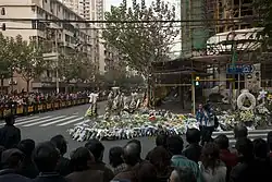 A large crowd stands behind metal fences around the base of a burned-out building, which is surrounded by many flowers and wreaths.
