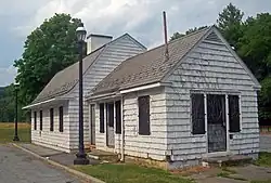 A small white building sided in shingles with a black asphalt bell-shaped roof, seen from its left. A slightly lower eing with a pointed roof projects toward the right. Its doors are screened and locked, and it looks neglected. Two black iron lamppots rise next to a curb and pavement at its left