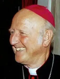 Colour three-quarter profile Press shot of a smiling, elderly bishop, with receding white hair, in a zucchetto and clerical collar.