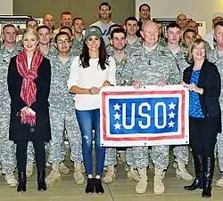 Dianna Agron and Meghan Markle, dressed casually and surrounded by military service personnel, hold a USO banner.