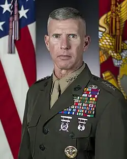 Caucasian male with high-and-tight haircut, graying hair, blue eyes; wearing Marine Corps service uniform with olive jacket and khaki shirt and tie; left chest full of military ribbons; shoulder epaulettes with four stars