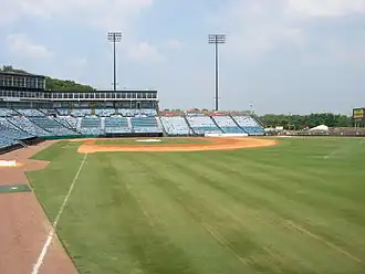 A view from the right field line of the seating bowl at Greer. Blue seats stretch from the right field wall, behind home plate, and beyond the third base dugout.