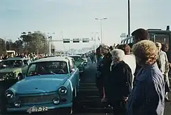 East German Trabant cars driving between dense crowds of people. Metal gantries over the road and a watchtower are visible in the background.