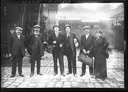 A group of five men and one woman, each carrying a travel bag, musette bag or parcel, cap or hat, some in ties, all in jackets.
