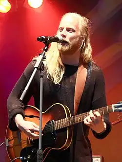 close-up of Högni Egilsson wearing a see-thru dark top, playing electric guitar and singing into a microphone onstage