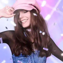 A woman with long brown hair smiles, eyes closed, wearing a light pink cap and a choker. She is dressed in a black mesh top under denim overalls, and confetti falls around her against a pastel purple and pink gradient.