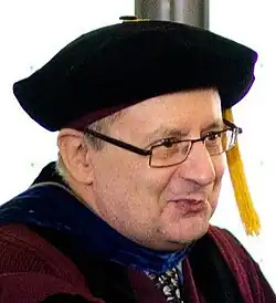 Three-quarter profile colour Press shot of a clean-shaven, smiling, bespectacled man wearing academic robes and hat.