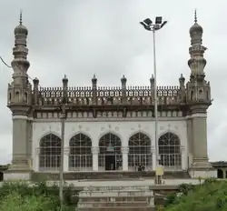 A mosque with five arched entrances, flanked by two minarets. The facade is white, and the minarets are brown. In the foreground are steps leading up to the mosque, with shrubs on either side.