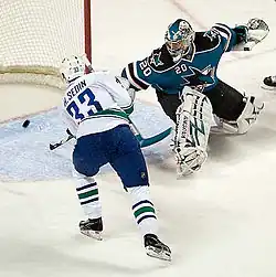 An ice hockey player wearing a white and blue jersey scores against a goaltender wearing a teal and black jersey with white pads. The player has both hands on his stick outstretched to direct the puck in the net, while the goaltender is off balance looking back at the puck in his net.