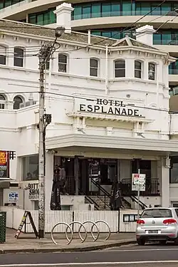 External shot of a two-storey building. The entrance is at left with the name displayed above. The building has a white picket fence in front. Two workmen are visible descending the entrance steps with a section of the same white fencing. To the right of the entrance is a hand-written notice in green-blue writing: "Patrons must be seated at all times". The building's façade is off-white.
