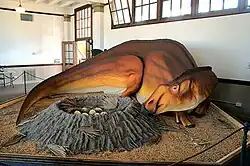 On a roped-off platform, an orange-gray dinosaur is curled around a gray crater of eggs.