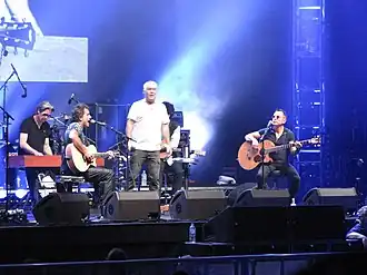 A stage shot with four men clearly visible. The man at left is leaning at a rectangular box; he has a microphone nearby. The second man is shown in right profile; he is playing a guitar and singing into a microphone while seated. The third man is standing in the middle of the stage with arms at his sides; he carries a microphone in his right hand. The fourth man is also seated while playing a guitar and at a microphone. A fifth man is obscured seated behind and to the right of the middle one. The men are surrounded by stage equipment including speakers, lights and additional microphones. A drum kit is partly visible but the drummer is obscured.
