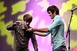 Ian Hecox standing to the left and Anthony Padilla standing to the right performing on stage at VidCon 2012, with Hecox pouring a bottle of water in a bucket held by Padilla