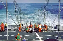 The rear deck of ship, with a large partially erect net visible near the center of the image. Many men in orange suits are working to free a white drone entangled in the net.