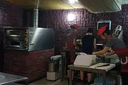 A dimly lit room with a pizza oven and a table where three employees stand with pizza ingredients.