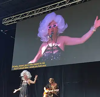 Bond, wearing a large white wig sings with Carerra, a guitar player, below a projected image of Bond