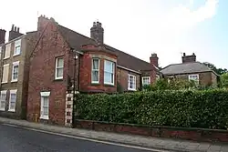 Large red brick house with projecting lower and upper bays, a high hedge screening the front garden and a blue plaque visible on side elevation