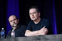 An American man with graying black hair sits behind a stand speaking to a crowd through a microphone.