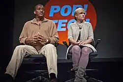 Ronald Cotton (left) and Jennifer Thomson-Cannino (right) sitting in two rolling office chairs in front of large red and blue Pop!Tech logo.