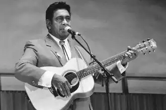 A man is shown in an upper body shot, he sings at a microphone while holding an acoustic guitar.