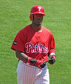 A dark-skinned man in a red baseball jersey, white baseball pants with red pinstripes, and a red batting helmet walks on a baseball field while taking off his black and red batting gloves.