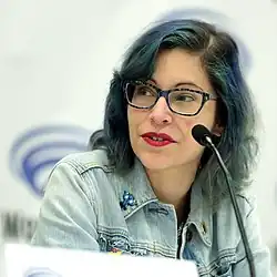 Photograph of Houser at a WonderCon panel with a microphone seen in front of her face as she looks slightly to her right. She is wearing a jeans jacket with pins, blue framed glasses, and her shoulder length hair is partly dyed blue.