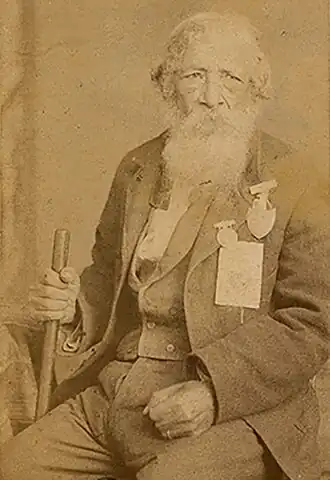 The picture is a sepia toned photograph that shows an African-American man in his eighties wearing a suit adorned with war medals and holding a cane.