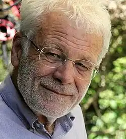 Colour Press-headshot of a smiling, elderly, white-haired, bespectacled man, with designer-stubble beard.