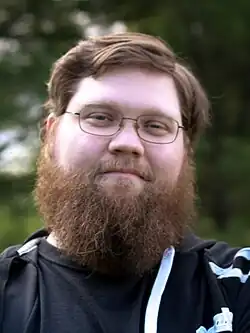 Justin Knapp—a Caucasian male with brown hair and a bushy beard—stands with his arms folded