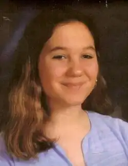 A smiling young adolescent girl with long, wavy brown hair and wearing a light blue striped shirt