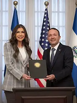 Kristi Noem and Gustavo Villatoro standing in front of U.S. and Salvadoran flags and holding a document