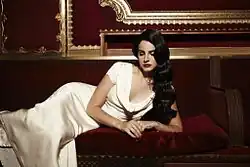 A woman wearing a white dress laying on a red velvet couch in front of a grandiose red wall with gold accents