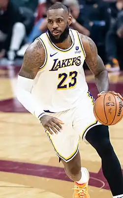 Full-body shot of LeBron James dribbling. He is a tall, black man with a buzz cut and a full beard. He is wearing a white jersey from the Los Angeles Lakers with the number 23.