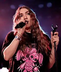 Lisa Heller singing on stage with the microphone in her right hand and the mic stand in her left. She is wearing a black shirt with pink stuffed rabbits.