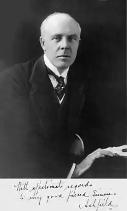 A balding, white-haired man wearing 1920s formal business attire (jacket and waistcoat with wing collar and tie) leans to his left on a table and looks directly at the viewer. His right hand rests in front of his left on the edge of the table next to some papers and a pen.