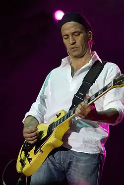 A full color picture of Urquijo holding a yellow electric guitar. He wears a black cap, white shirt, and blue jeans.