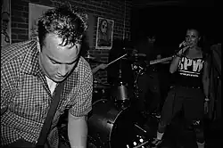 Foreground: Sweaty, make guitarist, and female singer, live on stage. In the background we see the bassist in the shadows, and hands of the drummer.