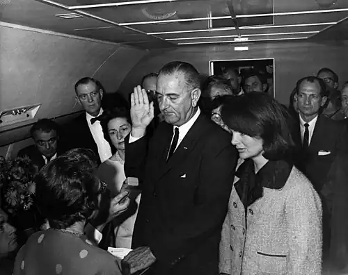 Lyndon B. Johnson raises his hand above an outstretched Bible as he is sworn in as President as Air Force One prepares to depart Love Field in Dallas. Jacqueline Kennedy, still in her blood-spattered clothes (not visible), looks on.