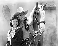 A black and white photograph of a man dressed as a cowboy with his right arm around a woman in a dress and his left hand holding the reigns of a horse.