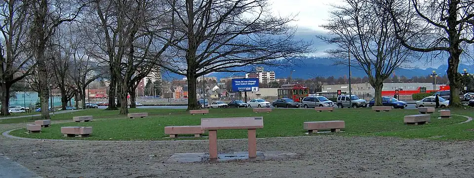 In a park, 14 coffin-like benches of pink stone are set in a circle. A higher slanted pink panel is visible in the foreground.