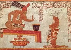 A man kneels before a man sitting on a platform. Between them sits a large container.