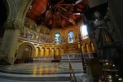 A view into the chancel is framed on the right by the lectern supported by a standing brass angel. The chancel is semi-circular and has a roof on wooden beams. The upper walls have brightly coloured mosaics of prophets and angels. The white marble altar and mosaic reredos of "The Last Supper" can be seen.