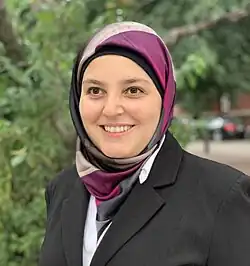 Portrait of Mona Minkara wearing a blazer and a patterned hijab, smiling in front of greenery.