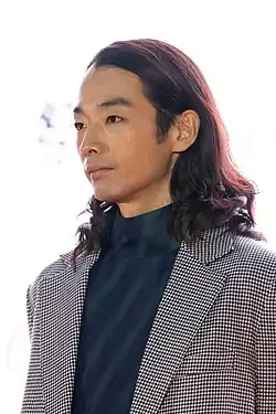 Moriyama at press event wearing suit and looking to left of camera