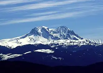 A snow-covered, conical mountain with two peaks rising over the foreground on a nearly clear day