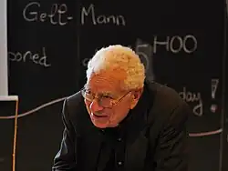 Murray Gell-Mann, speaking at a lectern in front of a chalkboard