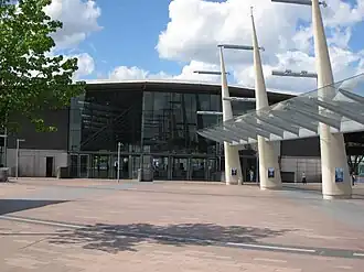 A grey building with grey lampposts in front and a green tree in the foreground all under a light blue sky with billowy, white clouds